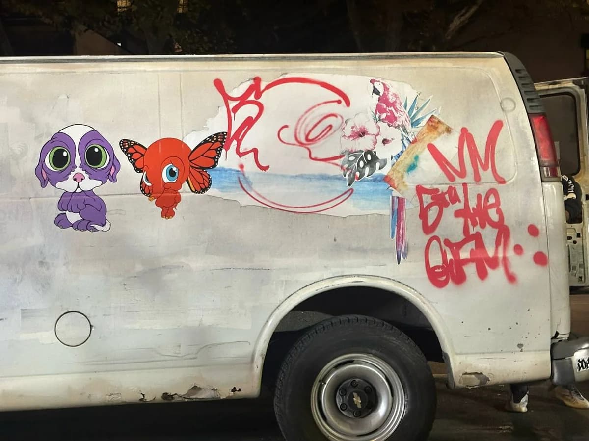 A white van features cartoon stickers alongside red spray-painted graffiti tags on its side.