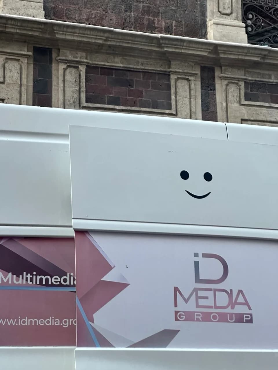 A simple black smiley face is drawn on a white panel above an ID Media Group sign in front of a stone building.